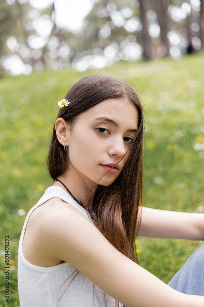 Portrait of brunette woman in top with daisy in hair looking at camera in park.