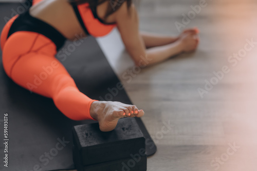 A young beautiful red-haired athlete is engaged in sports, doing stretching exercises at home on a sports mat with blocks. She is wearing an orange tracksuit. Close-up.