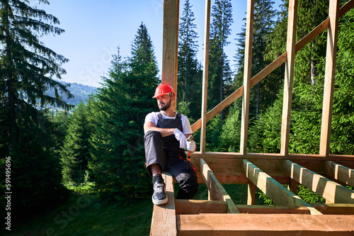 Carpenter constructing wooden frame two-story house near the forest. Bearded man holding hammer, dressed in protective helmet and overalls. Concept of ecological modern construction.