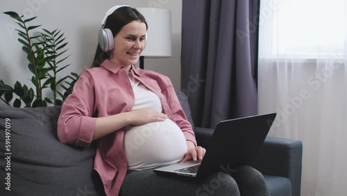 Happy pregnant woman in headphones sitting on sofa using laptop having video call conversation to family. Future mom talk to obstetrician-gynecologist, receive professional medical online consultation photo