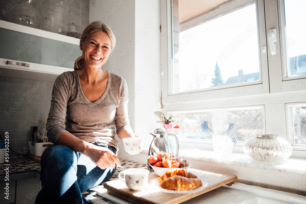 Happy woman having breakfast sitting on kitchen counter near window Happy woman having breakfast sitting on kitchen counter near window