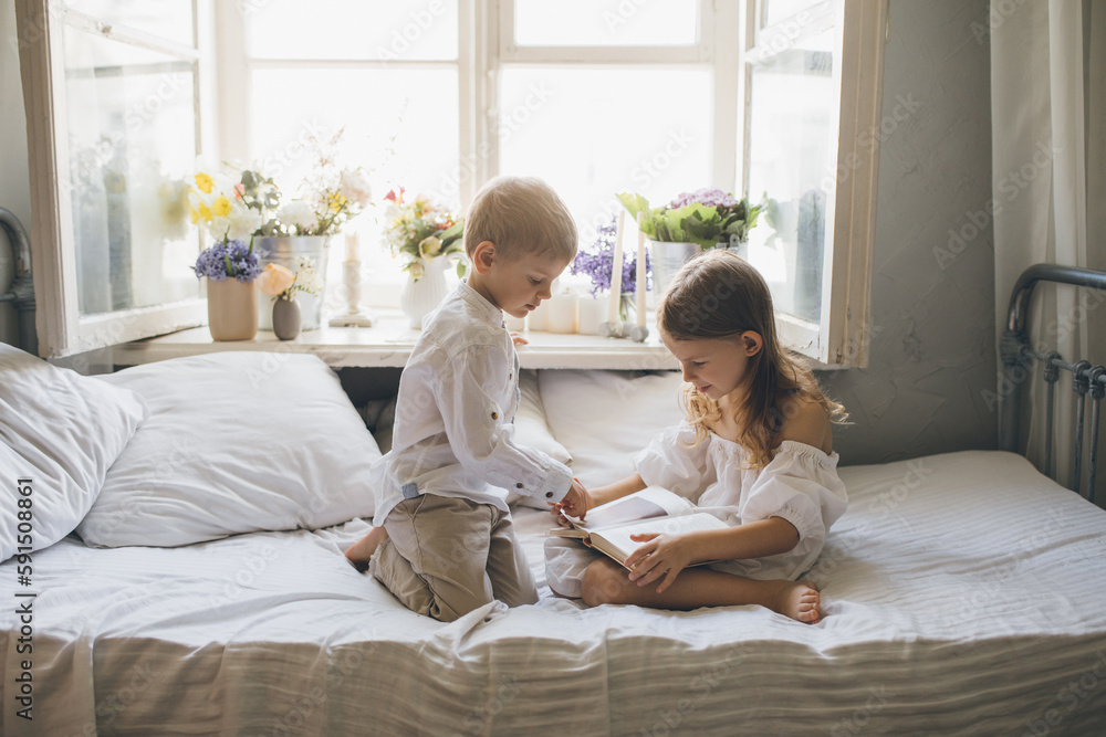 Cute boy and girl sitting on bed reading book together near window. Cute boy and girl sitting on bed reading book together near window.