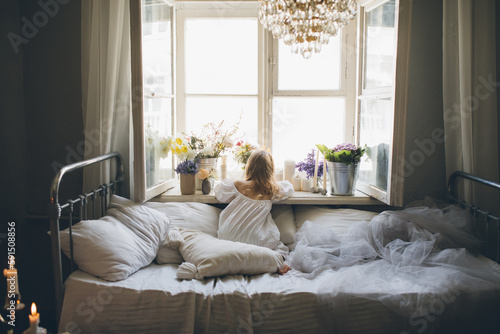 Girl in white dress sitting on a bed near window with lots of flowers.