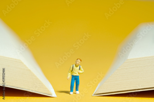 Miniature tiny people toy photography. A boy with school backpack standing in between two opened book page. Isolated on yellow background.