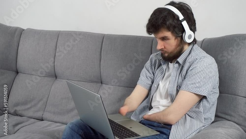 Disabled man with amputated two stump hands in headphones is listening music on laptop at home sitting on sofa. Problem of adaptation to life people with disabilities. Independent Invalid person.