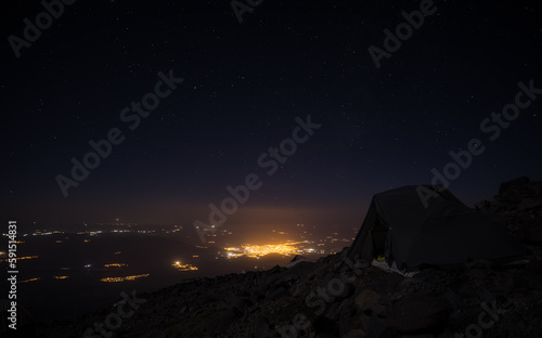 Night landscape in the mountains and a tent on the slope of Mount Ararat, against the backdrop of the glowing city of Dogubayazit and bright stars