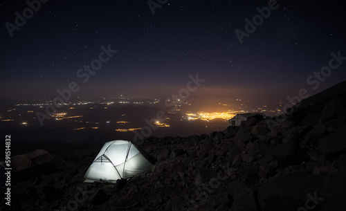 Night landscape in the mountains and a tent on the slope of Mount Ararat, against the backdrop of the glowing city of Dogubayazit and bright stars