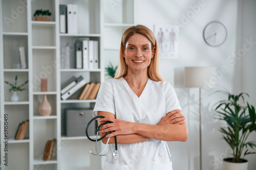 Holding stethoscope. Young female doctor in white coat is indoors