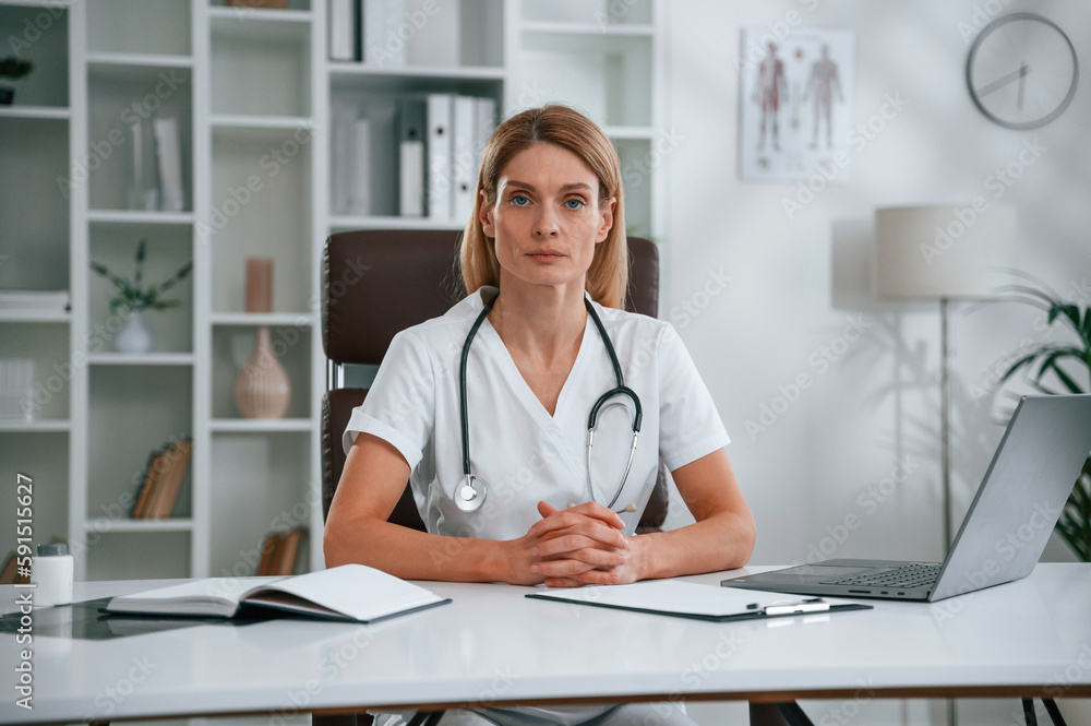 Sitting by the table with laptop. Young female doctor in white coat is indoors Sitting by the table with laptop. Young female doctor in white coat is indoors