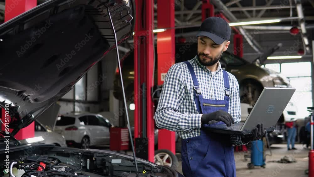 Experienced mechanic in uniform is using a laptop while repairing car in auto service Experienced mechanic in uniform is using a laptop while repairing car in auto service