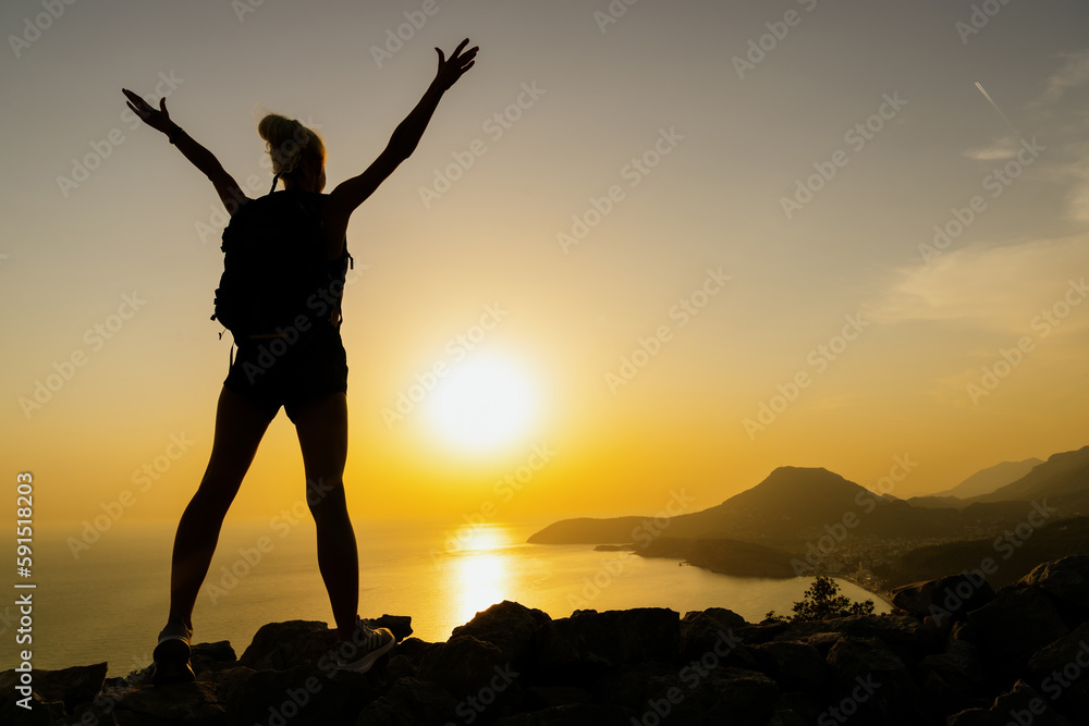 The silhouette of a joyful woman with her hands raised while hiking at sunset. Beautiful landscape