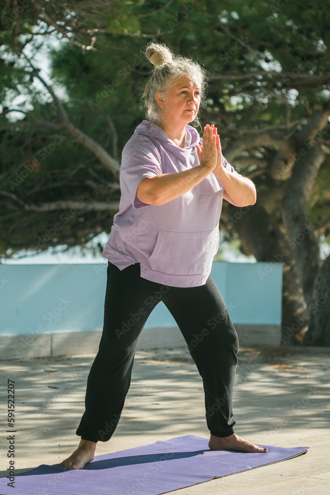 Serious aged woman in with dreadlocks hairstyle standing on beach against sea while practicing pose or asana during sunny day