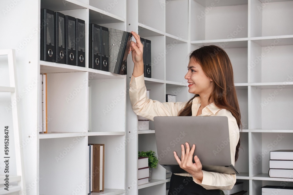 Smiling business woman taking binders from the shelf while using laptop computer at office Smiling business woman taking binders from the shelf while using laptop computer at office