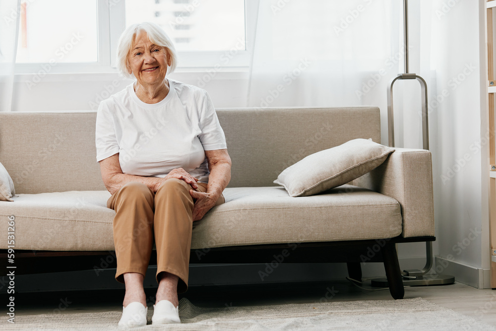 elderly woman sits on a sofa at home against the backdrop of a window and a happiness smile, stylish interior. Lifestyle retirement.