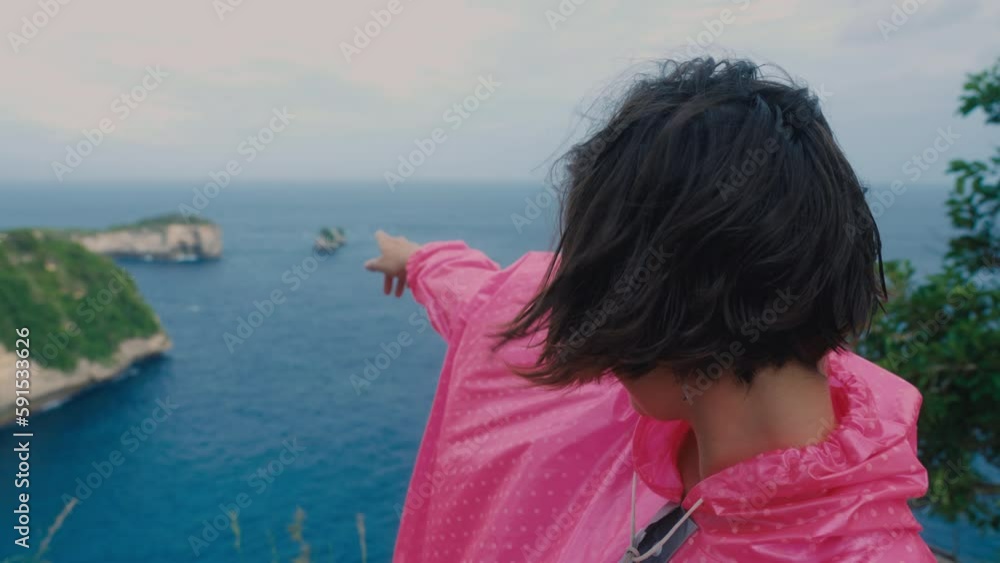 A cute Asian woman with short trendy hair standing on edge of cliff above blue ocean and pointing finger on distant island and land. Travel trip for happy female on seaside. Slow motion wind on coast.