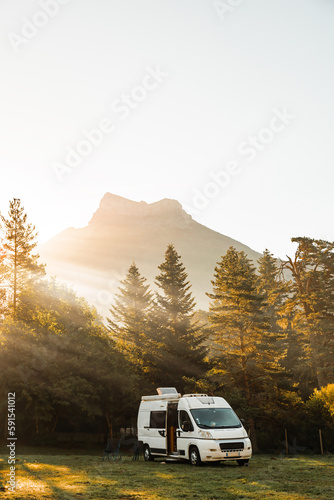 Camper van in a valley with amazing landscape views of forest, mountains and sun rays during sunrise. Van road trip holiday and outdoor summer adventure. Nomad lifestyle concept