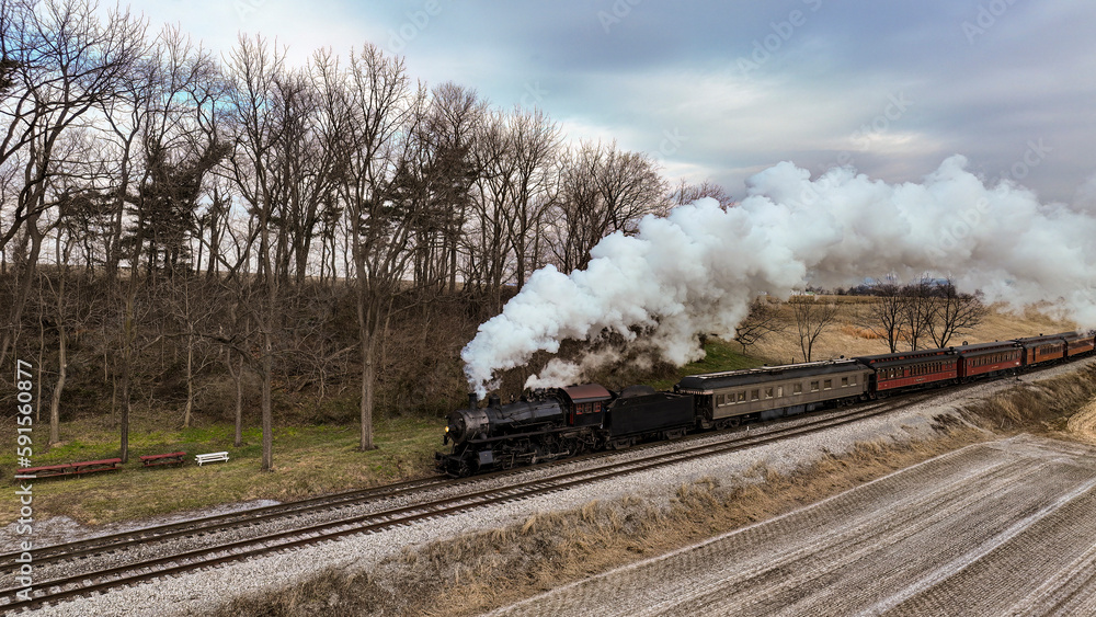 Obraz premium A Drone View of a Steam Locomotive Approaching Traveling Thru Fields and Meadows, Blowing White Smoke on a Winter Day