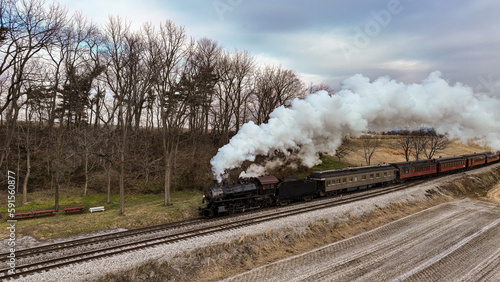 A Drone View of a Steam Locomotive Approaching Traveling Thru Fields and Meadows, Blowing White Smoke on a Winter Day