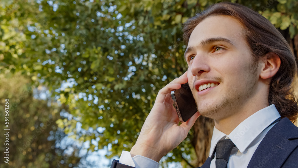 Portrait of the Businessman in a business suit in the park with mobile phone. Smiling businessman talking on a cell phone., outdoors. Young man in a business suit talking on a smartphone in nature Portrait of the Businessman in a business suit in the park with mobile phone. Smiling businessman talking on a cell phone., outdoors. Young man in a business suit talking on a smartphone in nature