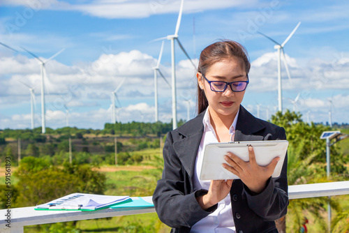 Energy engineer female working at wind farm. asian business woman wearing suit holding tablet and clipboard with windmill environment renewable energy.