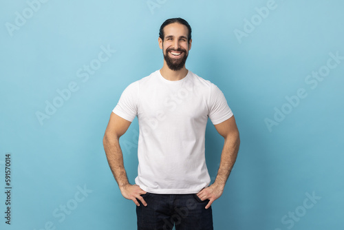 Portrait of bearded handsome man wearing white T-shirt standing with hands on hips, looking at camera with satisfied face and smiling. Indoor studio shot isolated on blue background.