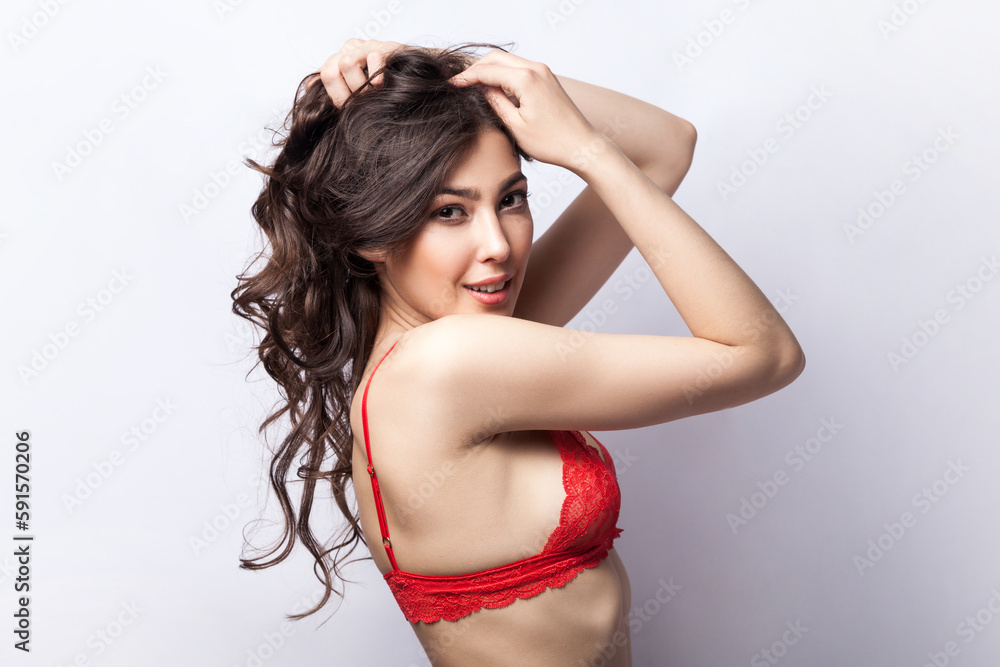 Side view portrait of brunette charming woman collecting her wavy hair and looking at camera with passion, wearing red underwear. Indoor studio shot isolated on gray background. Side view portrait of brunette charming woman collecting her wavy hair and looking at camera with passion, wearing red underwear. Indoor studio shot isolated on gray background.