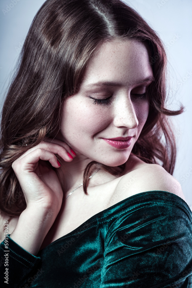 Naklejka premium Beauty portrait of pleased smiling beautiful woman with closed eyes wearing green dress, keeps hand near her face, having gently look. Indoor studio shot isolated on gray background.
