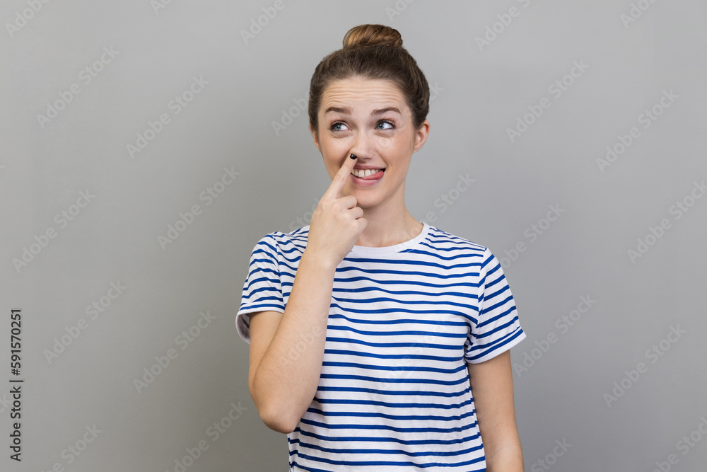 Portrait of childish woman wearing striped T-shirt sticking out tongue while picking nose, looking away with foolish silly expression. Indoor studio shot isolated on gray background. Portrait of childish woman wearing striped T-shirt sticking out tongue while picking nose, looking away with foolish silly expression. Indoor studio shot isolated on gray background.