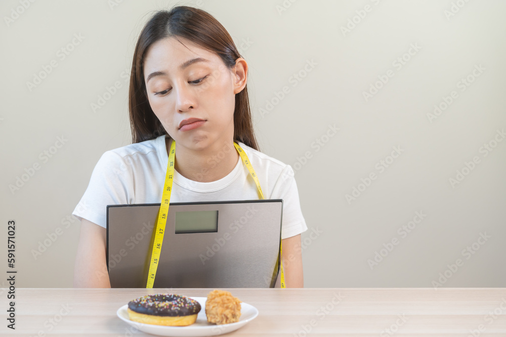 Diet, attractive asian young woman, girl restrained to eat doughnut, bakery and fried chicken, fast food to lose, loss weight, hugging weight scales on table at home. Passion, temptation when hungry. Diet, attractive asian young woman, girl restrained to eat doughnut, bakery and fried chicken, fast food to lose, loss weight, hugging weight scales on table at home. Passion, temptation when hungry.
