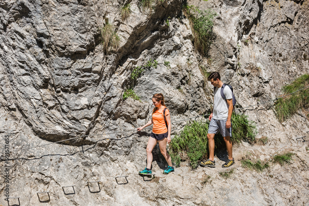 Man and woman with backpacks hiking carefully on the narrow, steep path above the rocky mountain river. Togetherness and outdoors pastime activity concept.