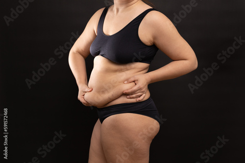 Overweight woman in black underwear massaging belly fat folds by hands, side view. Flaunt figure imperfections, cellulite body. Studio portrait isolated on black. Body positive, plus size people.