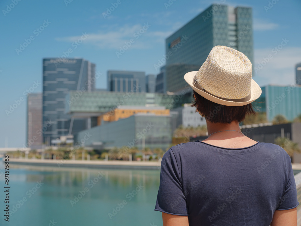 travel to the United Arab Emirates, Happy young asian female traveler in dress and hat look at View of Abu Dhabi Skyline in Al Maryah Island. Vacation and tourist destination concept.