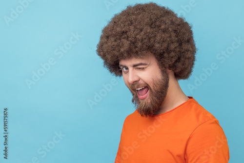 Portrait of flirting man with Afro hairstyle wearing orange T-shirt in good mood, smiling broadly and winking at camera with toothy smile. Indoor studio shot isolated on blue background. photo