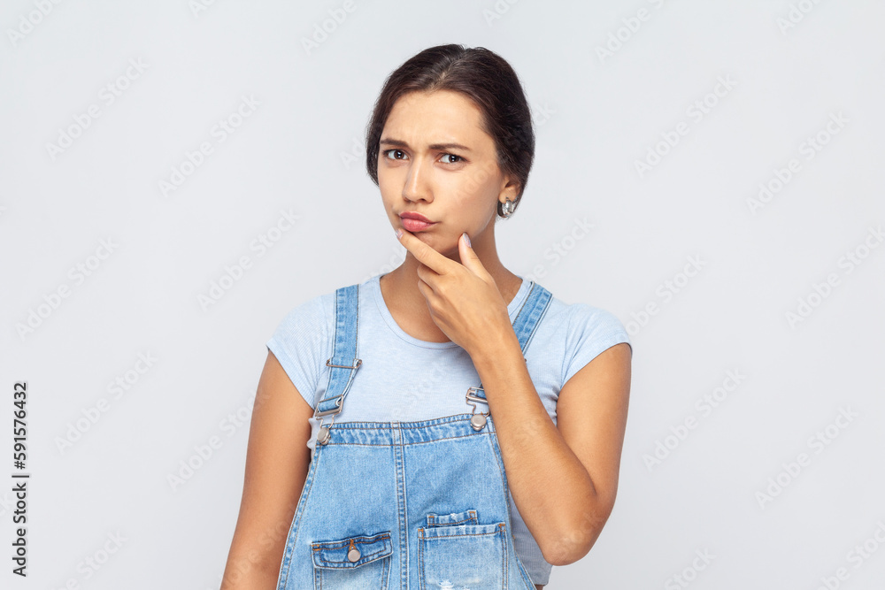 Portrait of thoughtful woman wearing denim overalls pondering serious issues, looking with uncertain hesitant expression, making difficult choice. Indoor studio shot isolated on gray background. Portrait of thoughtful woman wearing denim overalls pondering serious issues, looking with uncertain hesitant expression, making difficult choice. Indoor studio shot isolated on gray background.