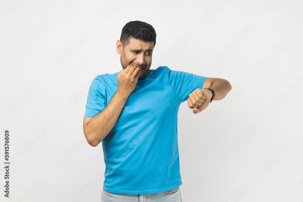 Portrait of nervous handsome unshaven man wearing blue T- shirt standing looking at smartwatch and rubbing his head, being late, biting fingernails. Indoor studio shot isolated on gray background. Portrait of nervous handsome unshaven man wearing blue T- shirt standing looking at smartwatch and rubbing his head, being late, biting fingernails. Indoor studio shot isolated on gray background.
