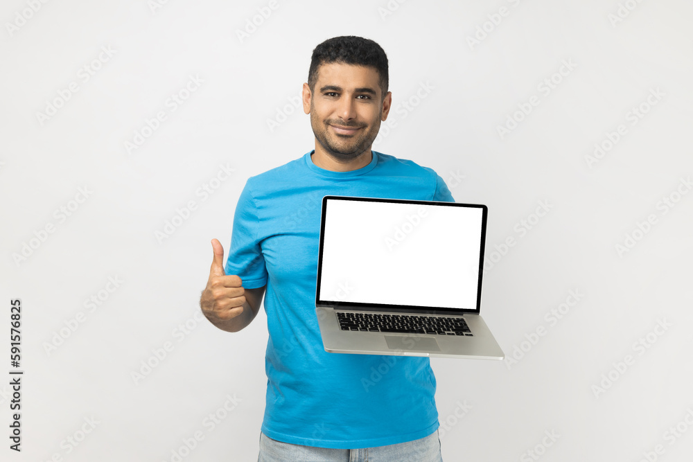 Portrait of delighted smiling attractive unshaven man wearing blue T- shirt standing showing blank screen of laptop, showing like gesture, thumb up. Indoor studio shot isolated on gray background. Portrait of delighted smiling attractive unshaven man wearing blue T- shirt standing showing blank screen of laptop, showing like gesture, thumb up. Indoor studio shot isolated on gray background.