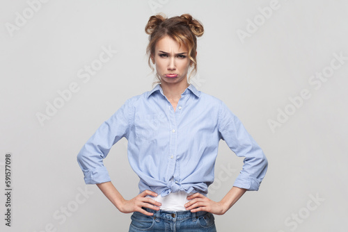 Portrait of blonde woman keeping hands on hips, has unhappy look, expressing regret and sadness, looking at camera with pout lips, wearing blue shirt. Indoor studio shot isolated on gray background.