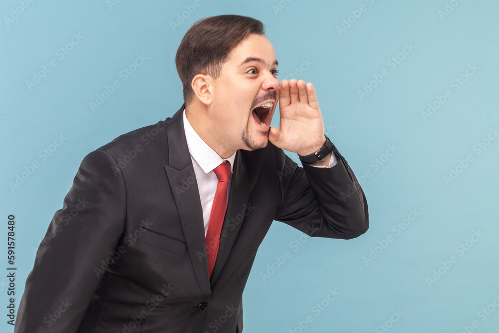 Side view of angry furious man with mustache standing standing and screaming loud, keeps hand near his mouth, wearing black suit with red tie. Indoor studio shot isolated on light blue background. Side view of angry furious man with mustache standing standing and screaming loud, keeps hand near his mouth, wearing black suit with red tie. Indoor studio shot isolated on light blue background.