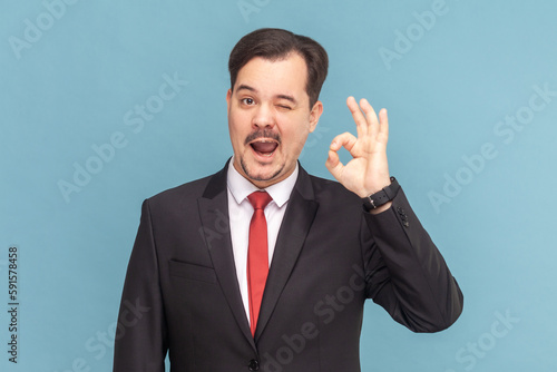 Portrait of funny optimistic man with mustache standing showing okay gesture, looking at camera and winking, wearing black suit with red tie. Indoor studio shot isolated on light blue background.