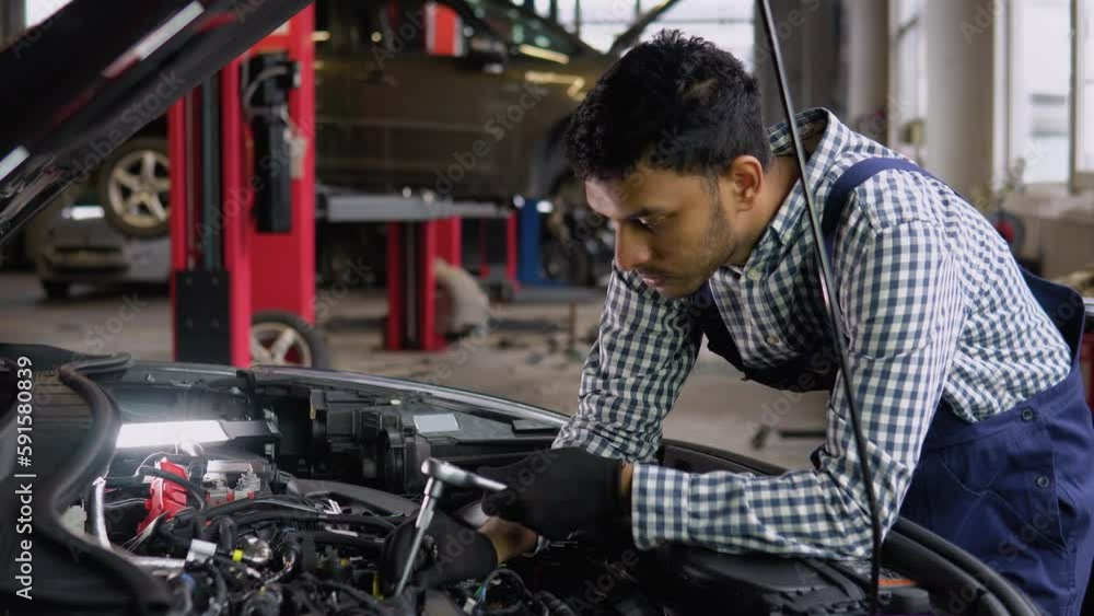 Indian auto mechanic examining broken car engine, standing near his car with open hood in a car repair shop Indian auto mechanic examining broken car engine, standing near his car with open hood in a car repair shop