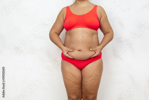 Overweight woman pinch fat folds of belly by hands, free copy space, white background. Bare woman in red underwear with cellulite body. Plus size people, body positive, feminism, self acceptance.