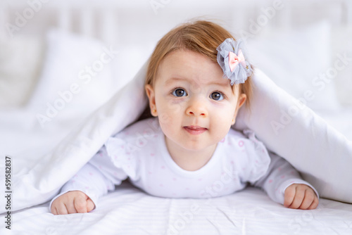 funny surprised baby girl looks out from under the blanket on the bed in white clothes with her mouth open, a small child on a cotton bed at home woke up in the morning after sleeping
