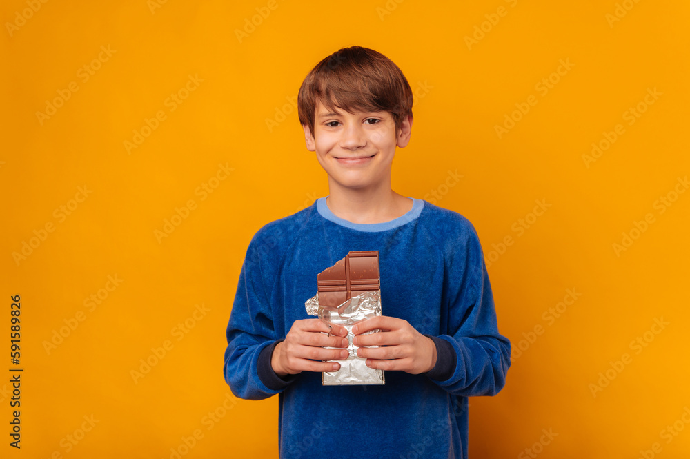 Handsome smiling young teen boy is holding a bitten bar of milk chocolate over yellow backdrop. Handsome smiling young teen boy is holding a bitten bar of milk chocolate over yellow backdrop.