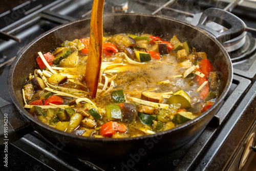 A wooden spatula mixes udon with stewed vegetables in spices in a frying pan.