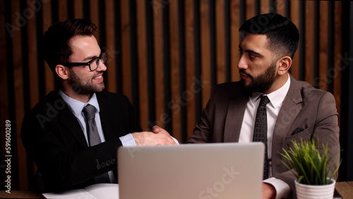 Two young men smile and shake hands as they meet at the table in the company office