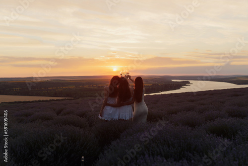 Young women enjoying view of sunset in the lavender field. They hold glasses of wine to celebrate their vacation. Copy space.