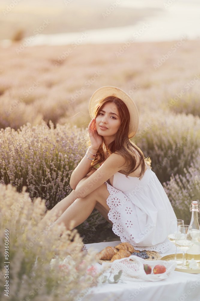 Young woman having a picnic on the lavender field in a nice sunny day. Female sitting on the picnic blanket and smiling.