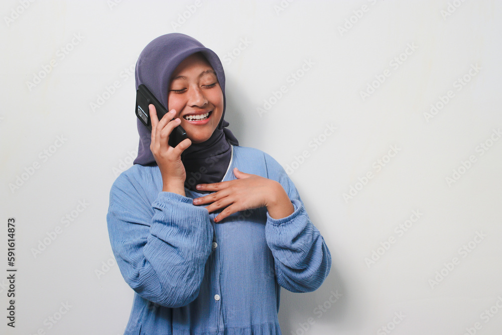 Happy young Asian girl in hijab touch her chest while listening her opponent to make a funny joke on a phone call Happy young Asian girl in hijab touch her chest while listening her opponent to make a funny joke on a phone call