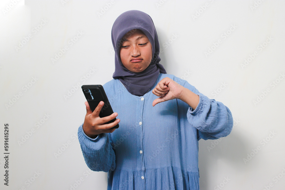 Young Asian girl in hijab doing thumb down while holding a smartphone Young Asian girl in hijab doing thumb down while holding a smartphone