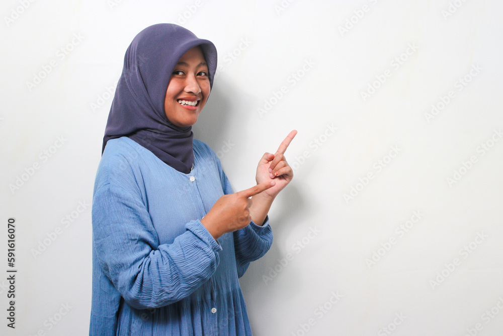 Excited young Asian girl in hijab pointing finger right on white background. Excited young Asian girl in hijab pointing finger right on white background.
